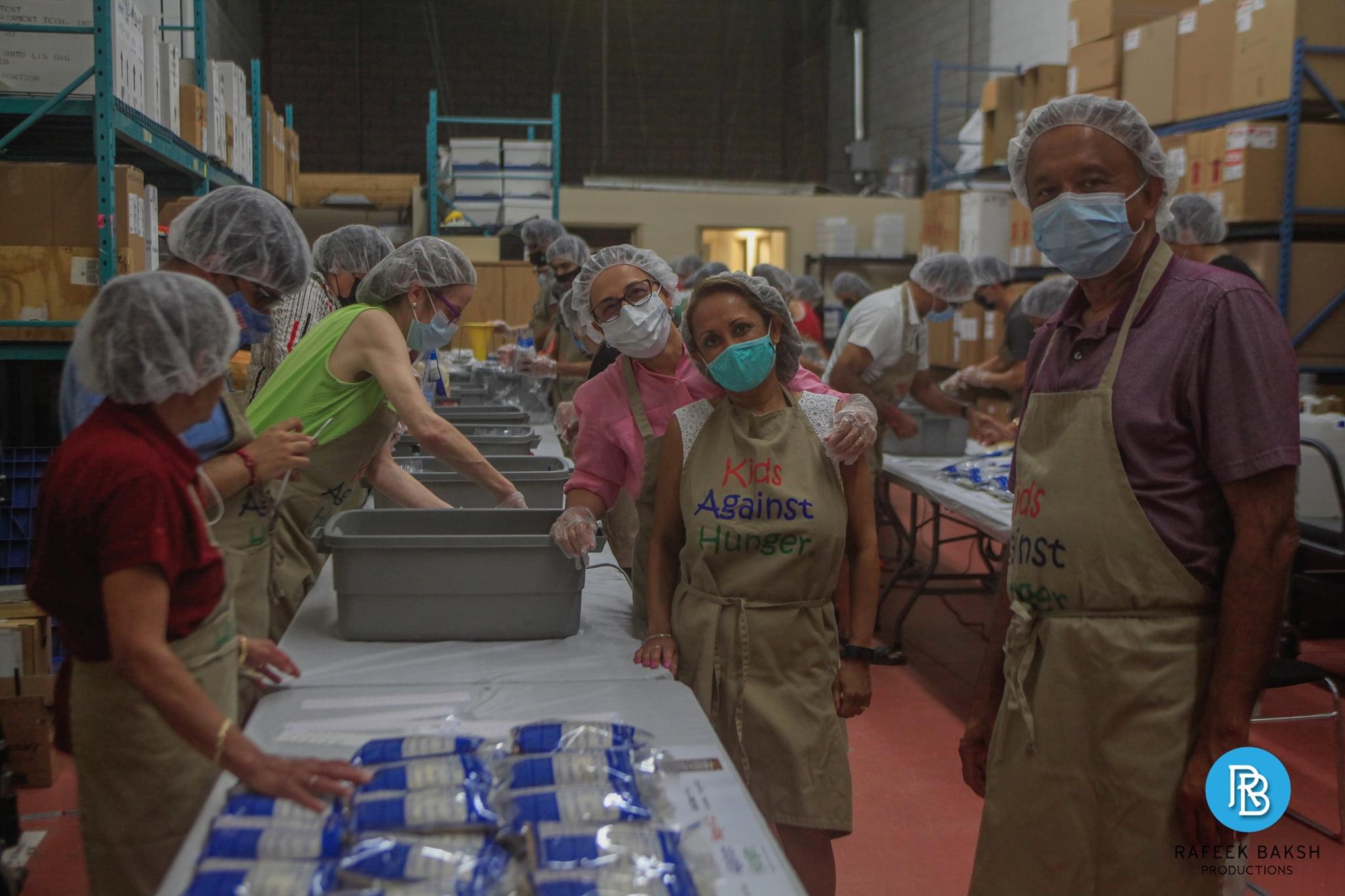 Volunteers standing along a table in a factory setting wearing masks while packaging meals for Kids Against Hunger Canada