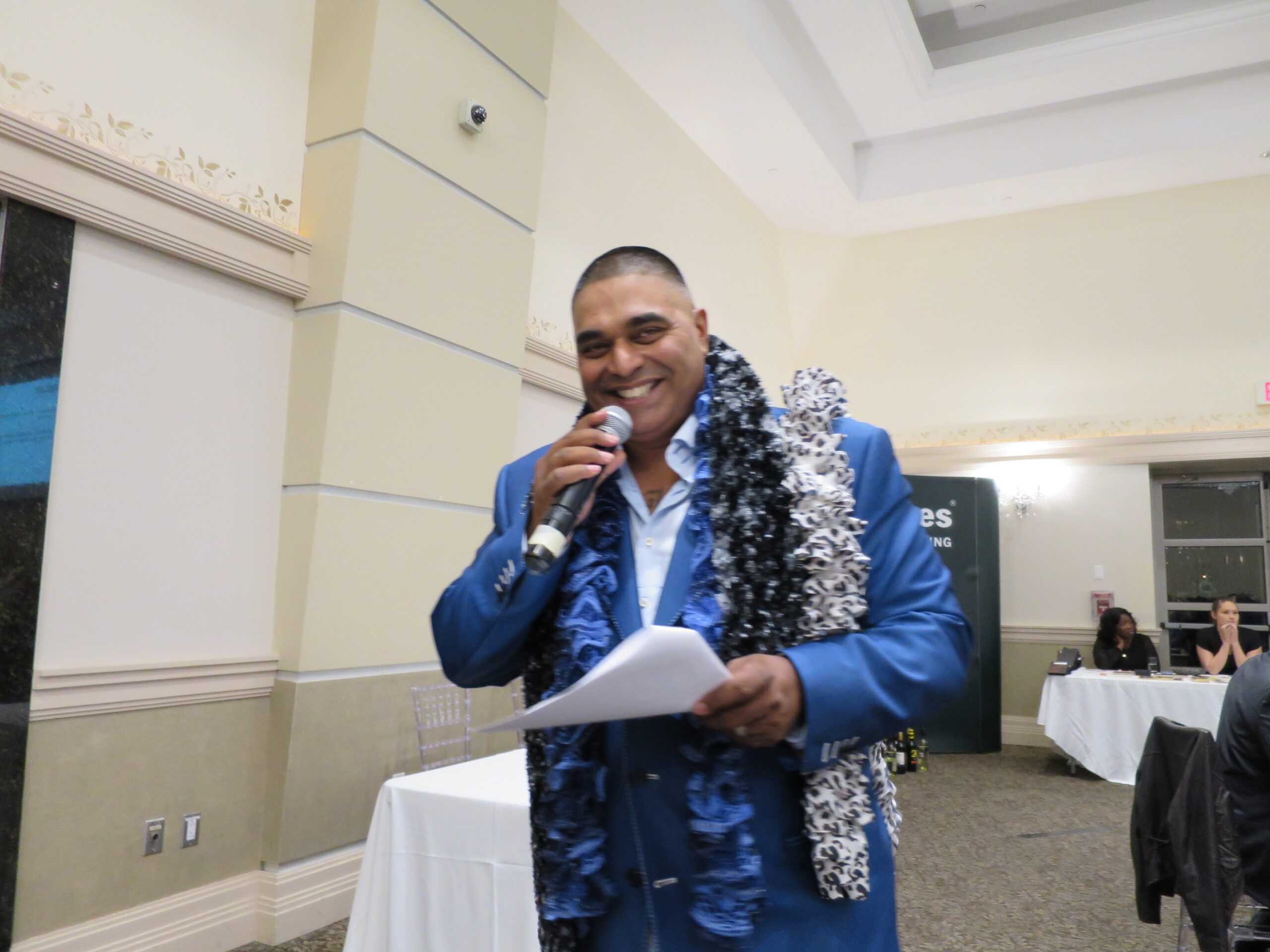 Keynote speaker holding a microphone and smiling at the 2015 Emajjin gala, wearing several scarves